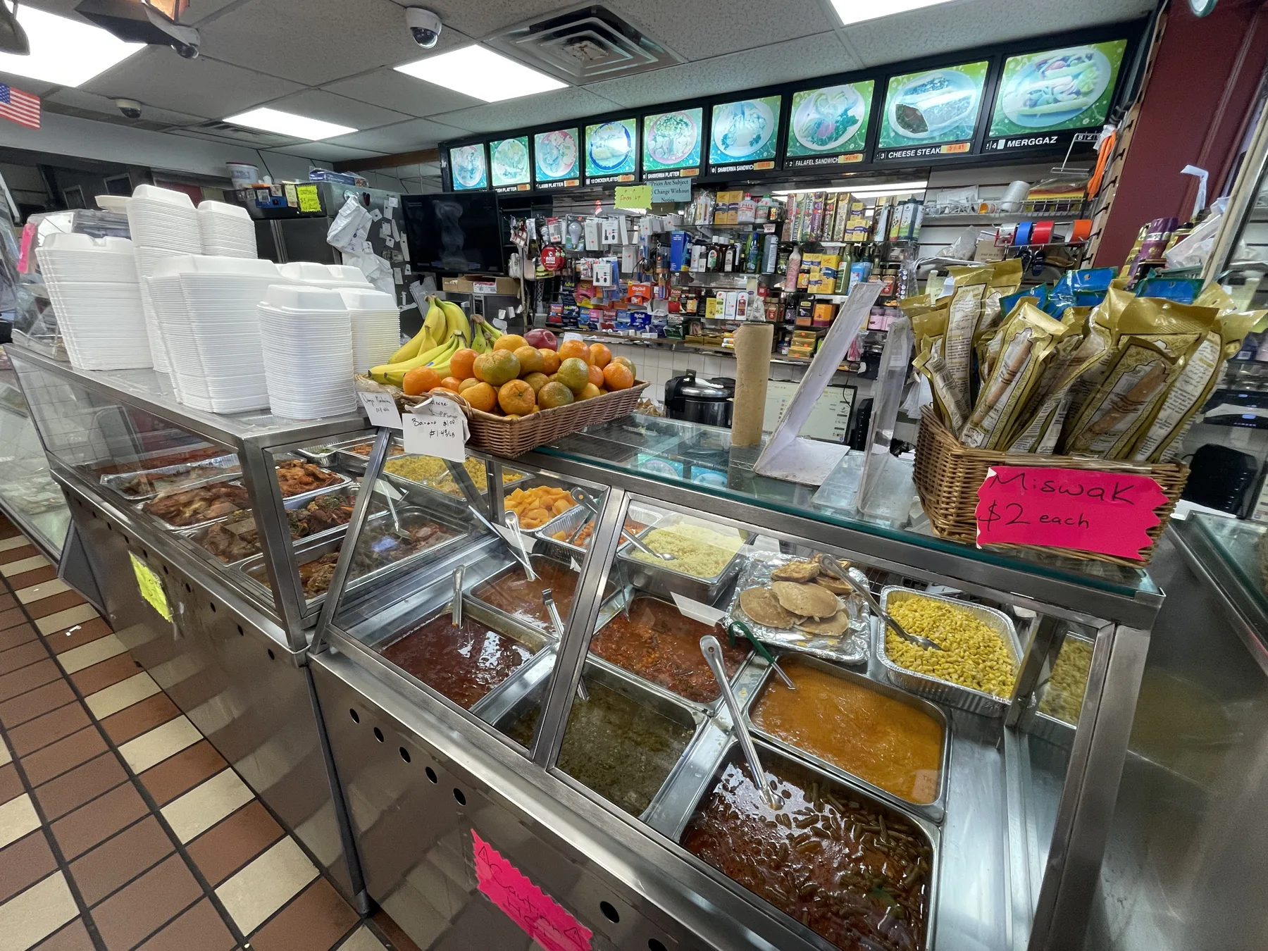 The full restaurant counter at Makkah Market — steam trays of cooked plates, the numbered menu board with shawarma, falafel and kebab platters, hanging fruit baskets and stacks of takeout boxes